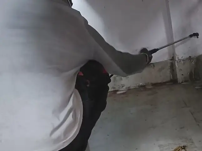 Technician spraying along a wall and floor edge under a bed frame