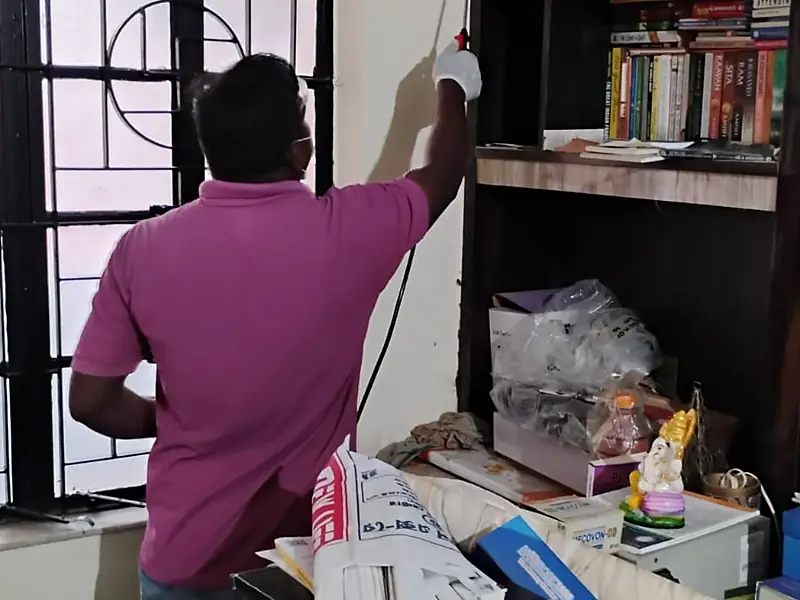 Technician spraying the upper corner of a wall beside a bookshelf in an apartment